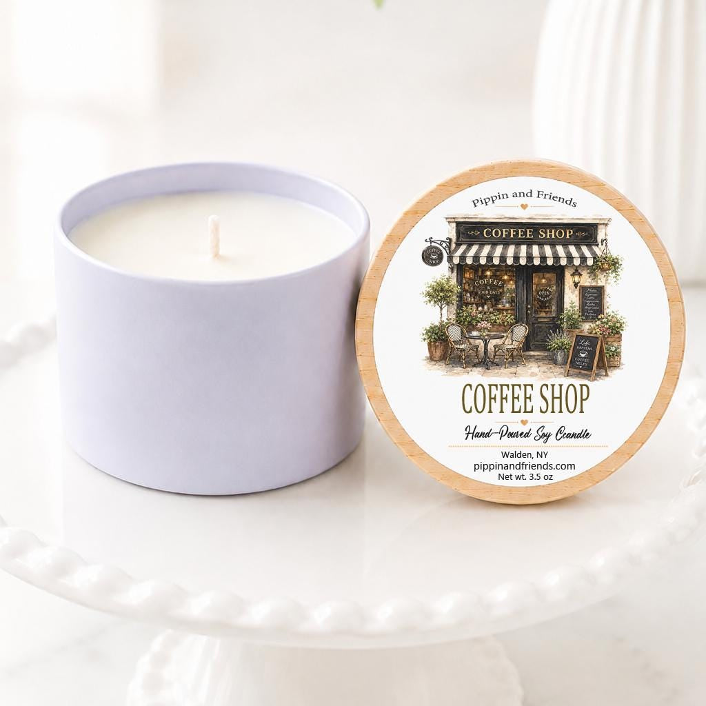 A soy candle in a matte cylinder container sits next to its round lid on a white pedestal. The lid features an illustration of a coffee shop and the text 'Coffee Shop'