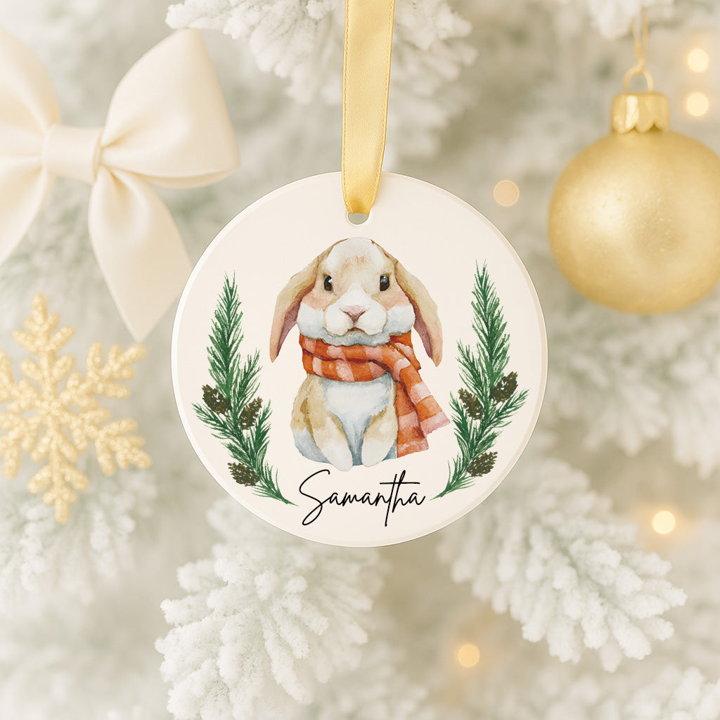 A white Christmas tree ornament featuring a rabbit wearing a red scarf and surrounded by pine branches and pine cones.