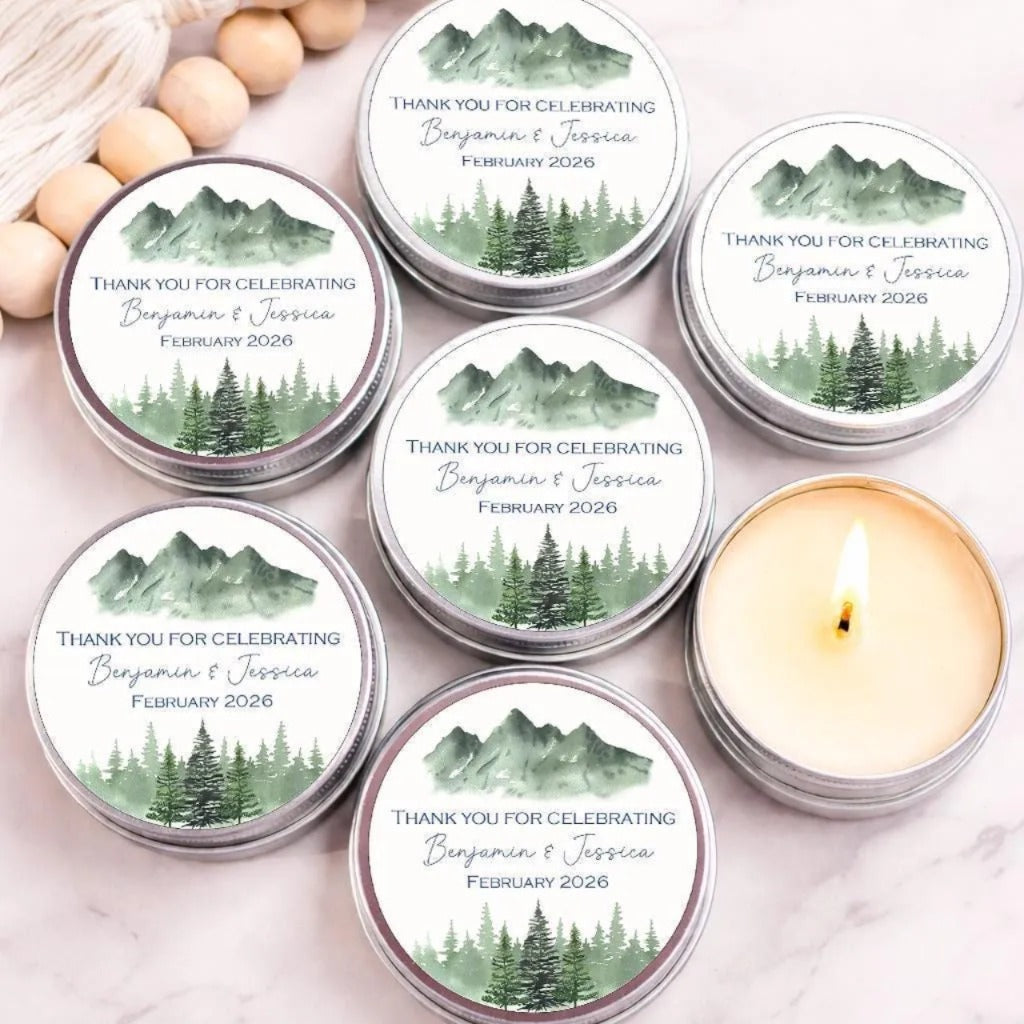 Personalized mountain wedding candle favors in silver tins featuring a watercolor mountain and pine forest design, displayed on a marble surface with one candle lit.