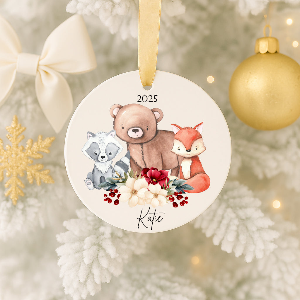 A white ornament with a bear, raccoon, and fox on it, surrounded by flowers and berries, hangs on a Christmas tree.