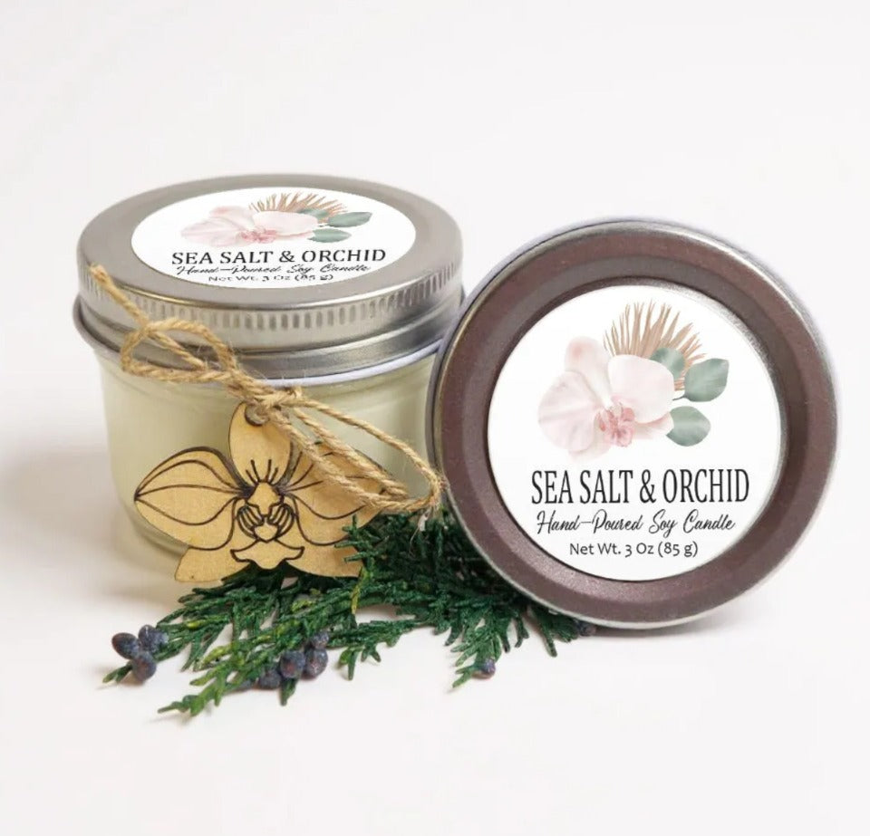 two jars of sea salt and orchid scented candles, with a sprig of greenery and a wooden ornament in the foreground.
