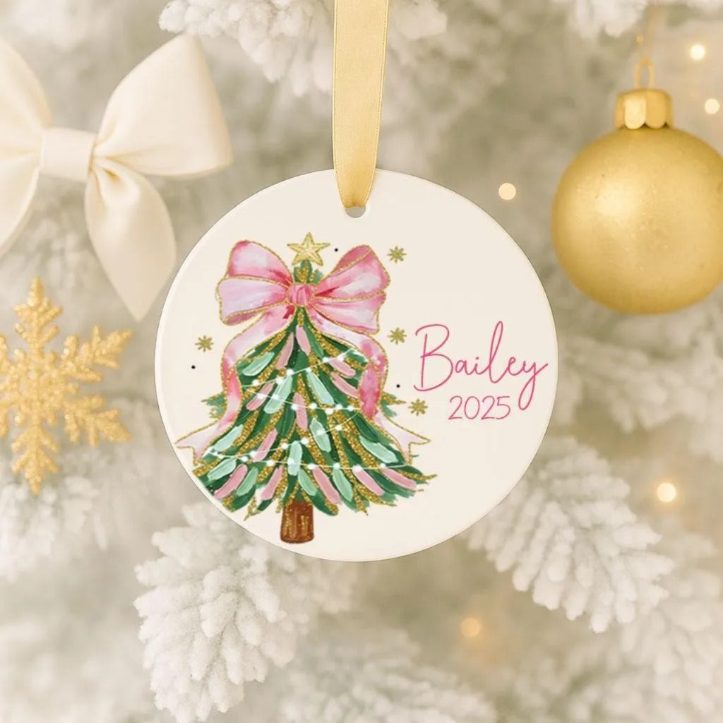 A white Christmas tree ornament with a pink bow and green and gold accents, hanging on a tree.