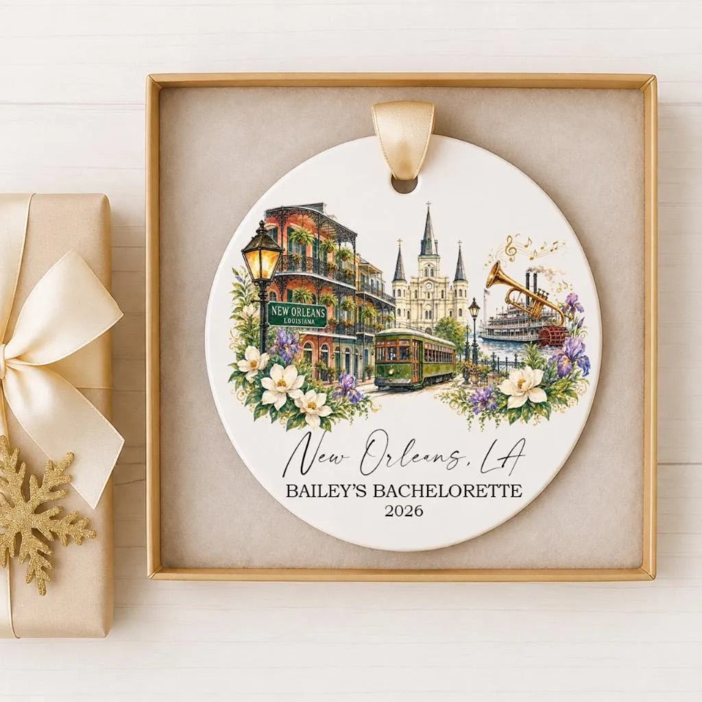 A decorative ornament featuring a painting of the New Orleans skyline, including the Cathedral of New Orleans, a trolley car, and a bouquet of flowers. The ornament is presented in a gold frame and is labeled as a "New Orleans, LA" bachelorette ornament from the year 2026.