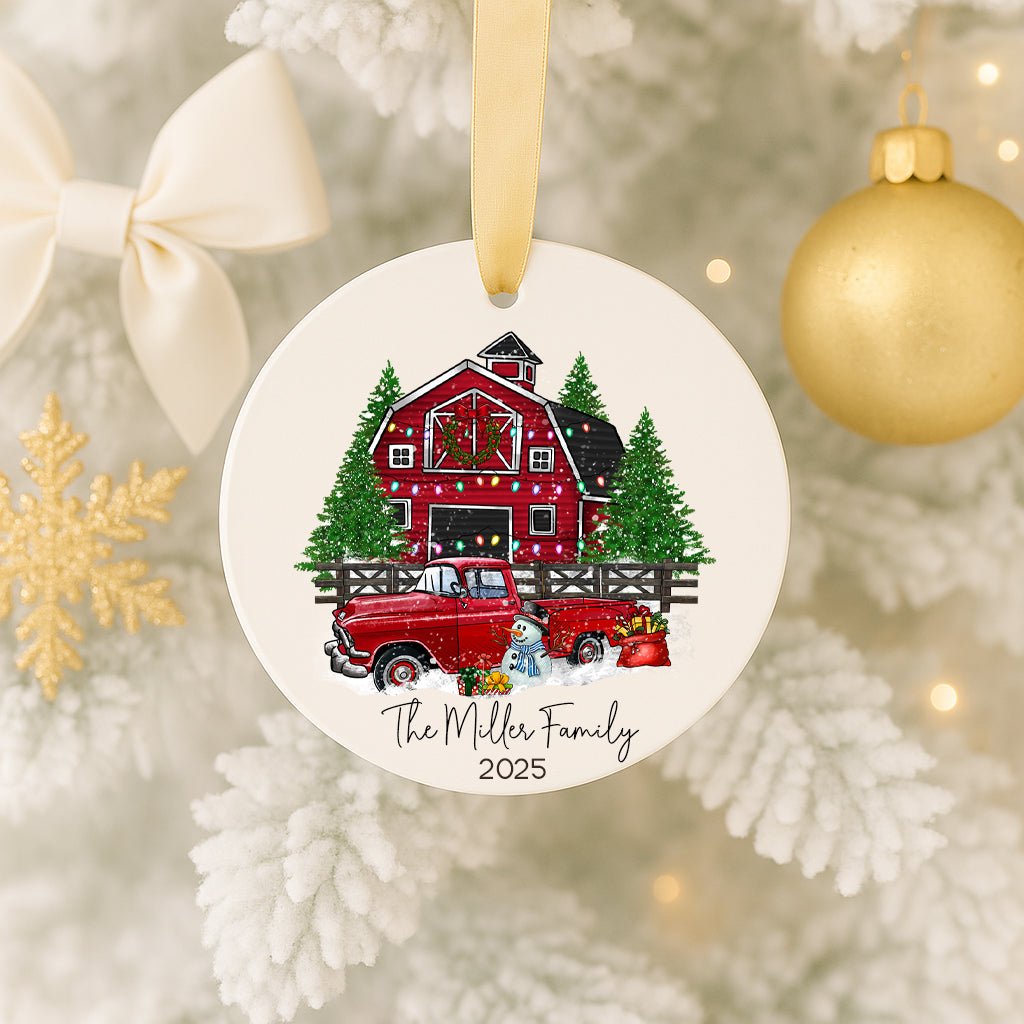 A white ornament with a red barn and Christmas trees, along with a red truck and a snowflake ornament, hanging on a Christmas tree.