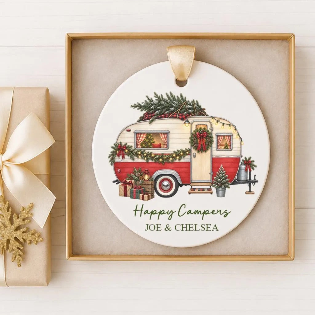 A white ceramic ornament with a red and white camper trailer decorated with Christmas lights and greenery, placed in a gold frame.