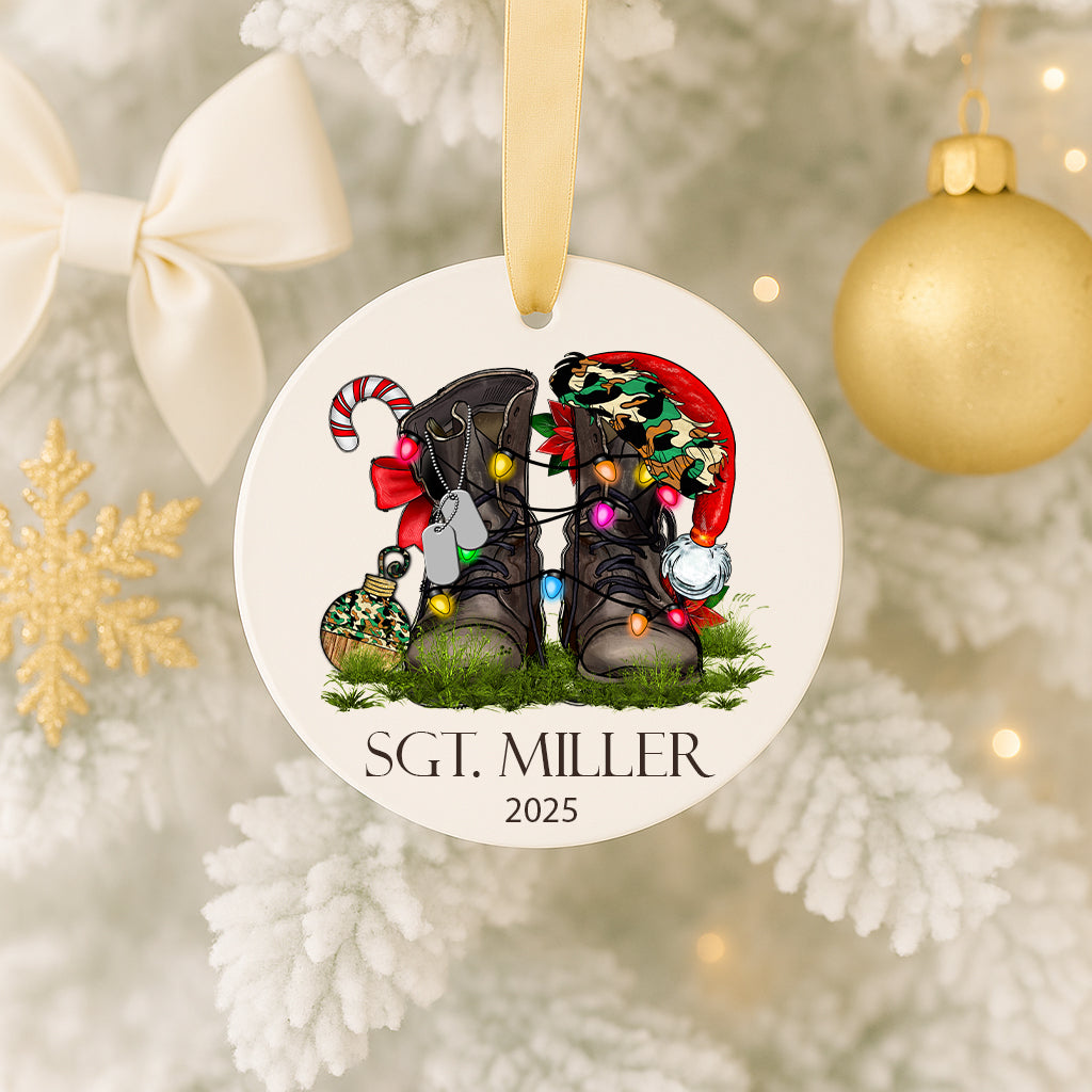 A white ornament with a Christmas tree and a pair of boots, along with a ribbon and a snowflake ornament, is hanging on a tree.