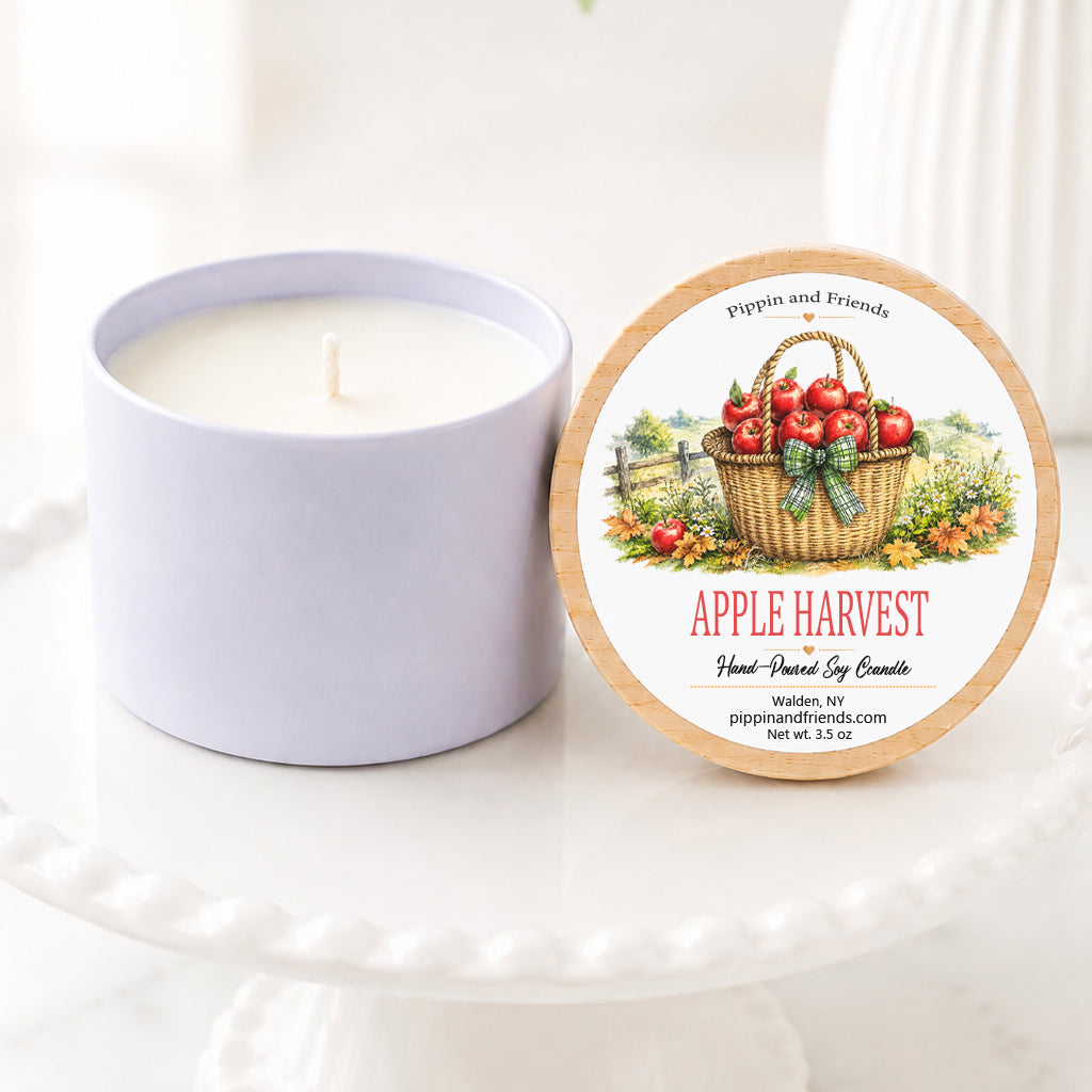 A soy candle in a matte container sits next to its round lid on a white pedestal. The lid features a vintage-style illustration of an apple basket with red apples and green leaves, with the scent name 'APPLE HARVEST' printed below.