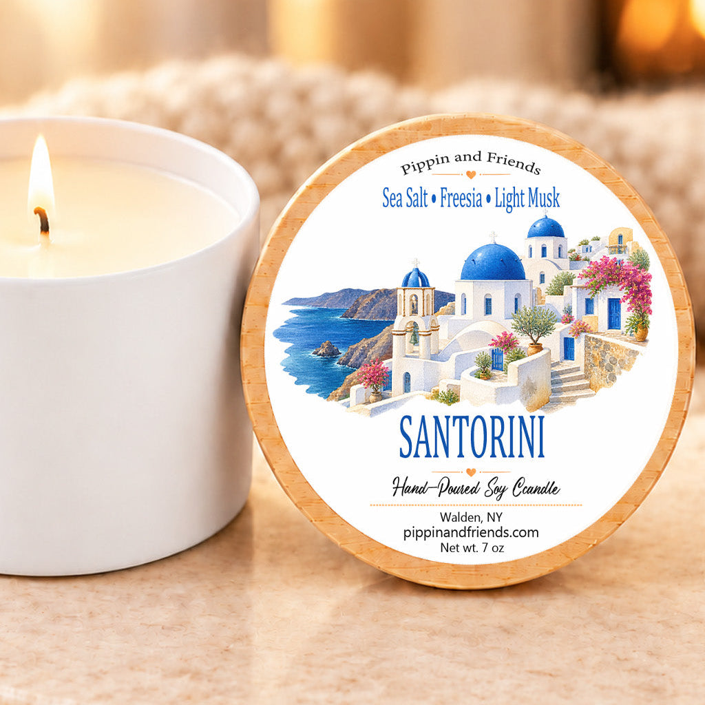 A round wooden-swatched candle tin labeled 'Santorini' by Pippin and Friends, shown beside a lit white candle on a warm beige stone surface with a softly blurred indoor background.