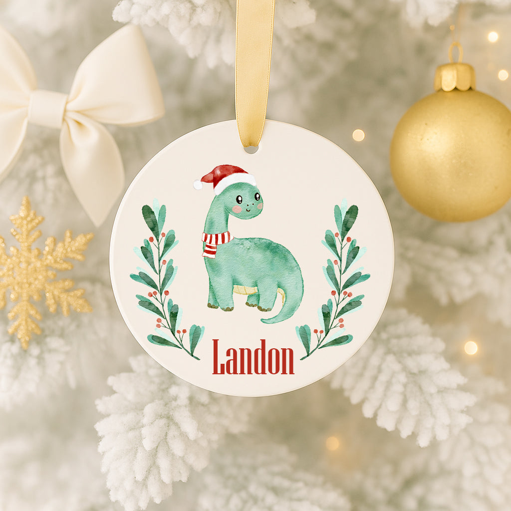 A white ornament with a green dinosaur wearing a Santa hat and a red scarf, surrounded by green leaves and red berries, hangs on a Christmas tree.