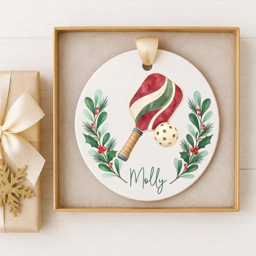 A ceramic ornament with a tennis racket and a ball, surrounded by a wreath of holly leaves and berries, with the name "Molly" written on it. The ornament is displayed in a gold frame, and there is a gift box with a ribbon visible in the background.