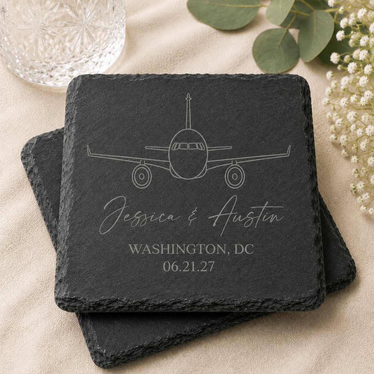 Personalized black slate coaster featuring a laser engraved airplane design with custom names, wedding location, and date. The natural-edge coaster is displayed in a stacked arrangement on a soft neutral background with greenery and crystal glassware, making it an elegant aviation-themed wedding favor.