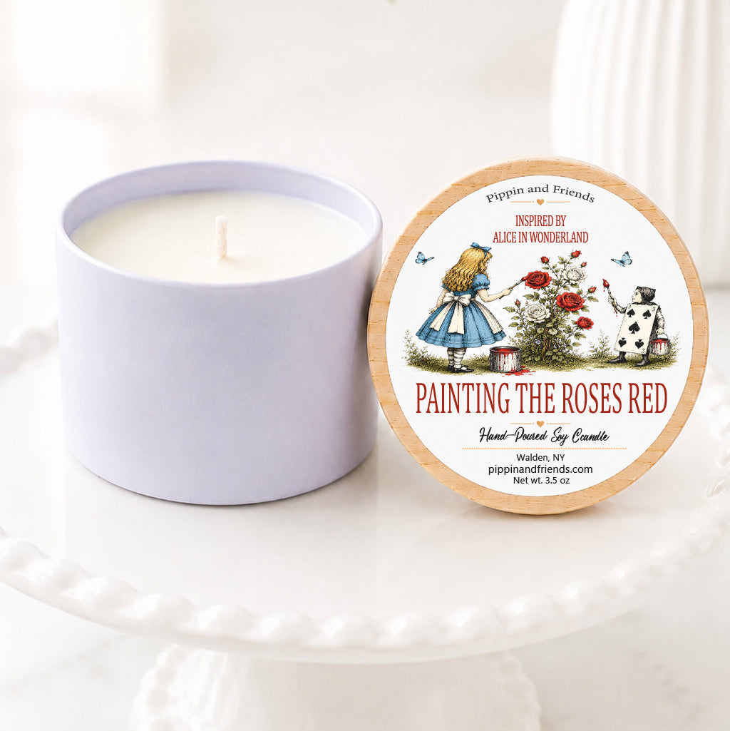A hand-poured soy candle sits on a white scalloped pedestal beside its matching wooden lid. The lid features an Alice in Wonderland–inspired label with a vintage-style illustration of Alice painting white roses red while the Two of Spades card gardener paints beside her. The label reads "Inspired by Alice in Wonderland – Painting the Roses Red" with "Hand-Poured Soy Candle" below. The candle is styled on a bright marble surface with a softly blurred white vase of flowers in the background, c