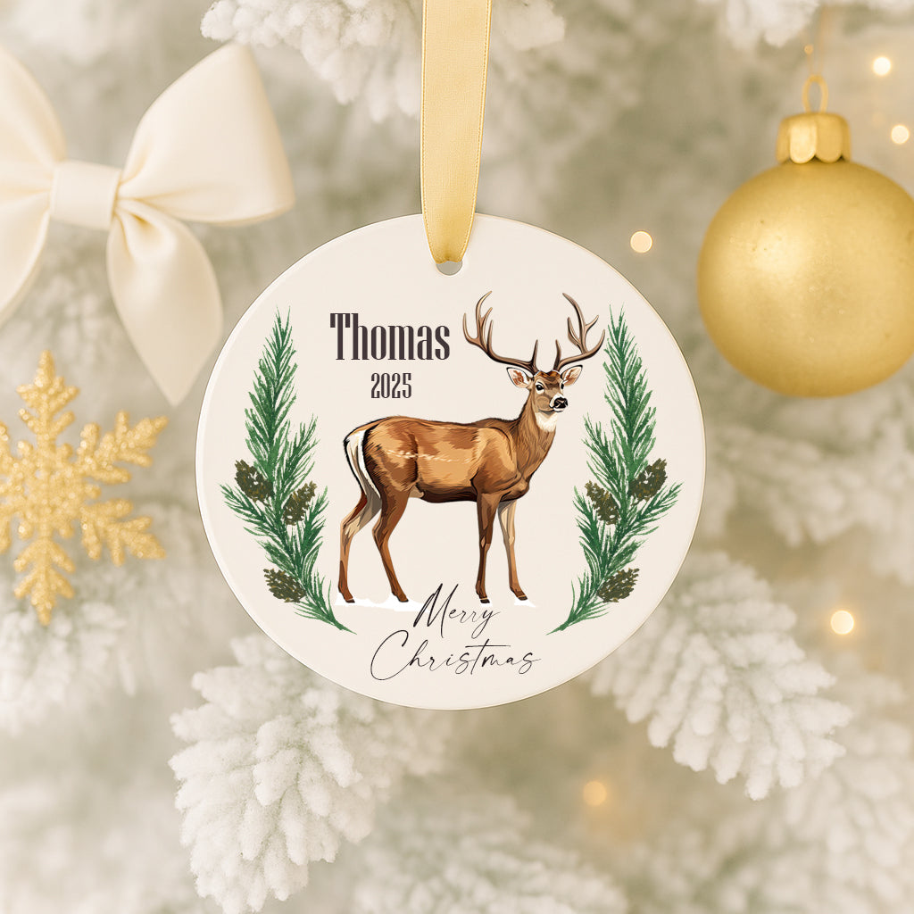 A white ornament with a deer and pine tree design hangs on a Christmas tree, surrounded by other ornaments and a gold ornament.
