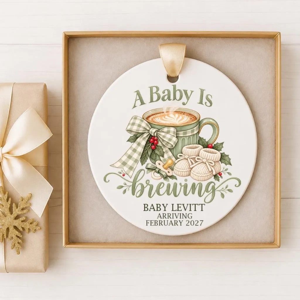 A decorative ornament with the text "A Baby Is Brewing" and "BABY LEVITT ARRIVING FEBRUARY 2027" on it, along with illustrations of a cup of coffee, a baby boot, and a bow.