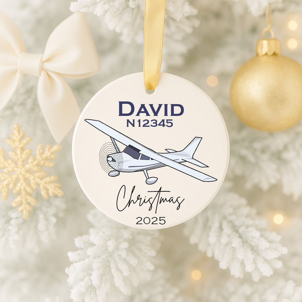 A white ornament with a blue airplane illustration and the name "DAVID" and the year "2025" printed on it, hanging on a Christmas tree.