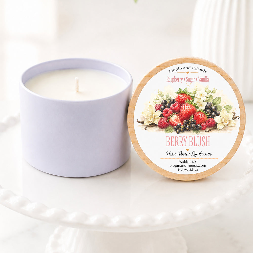 A soy jar candle next to its round lid, which features a berry-themed illustration and reads 'Berry Blush Hand Scented Soy Candle' by peppermint and friends. The candle and lid are displayed on a white ceramic pedestal with a soft-focus background of green leaves and white flowers.