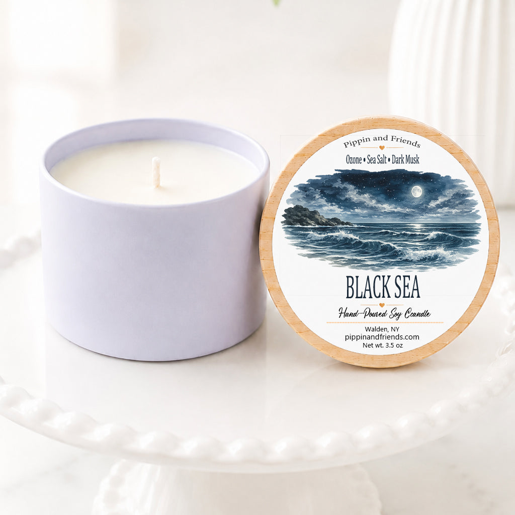 A soy candle in a matte cylinder container sits next to its round lid on a white pedestal. The lid features a natural wood rim and a label with a blue and black ocean illustration and the text 'BLACK SEA' and 'Hand Poured Soy Candle.