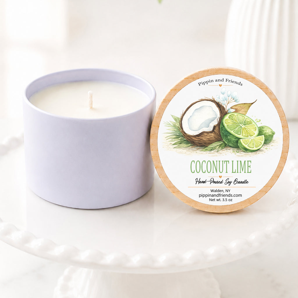 A soy candle in a matte cup container sits next to its round lid on a white pedestal. The lid features a natural wood border and an illustration of a coconut and lime slices with the text 'COCONUT LIME' and 'pippin and friends'.