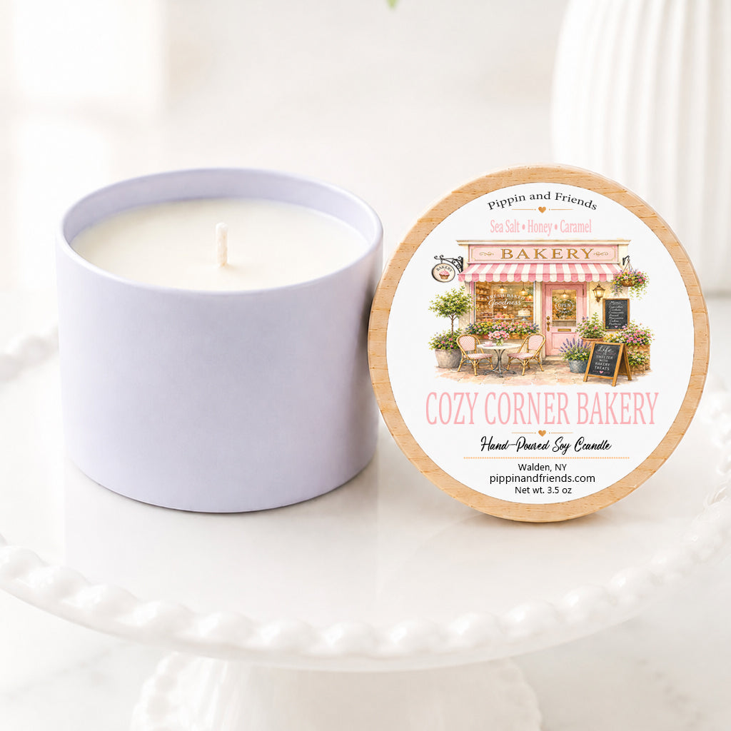 A soy candle in a matching cylindrical container sits beside its round lid on a white pedestal. The lid features a bakery-themed illustration and the scent name 'Cozy Corner Bakery' by Peppin and Friends.
