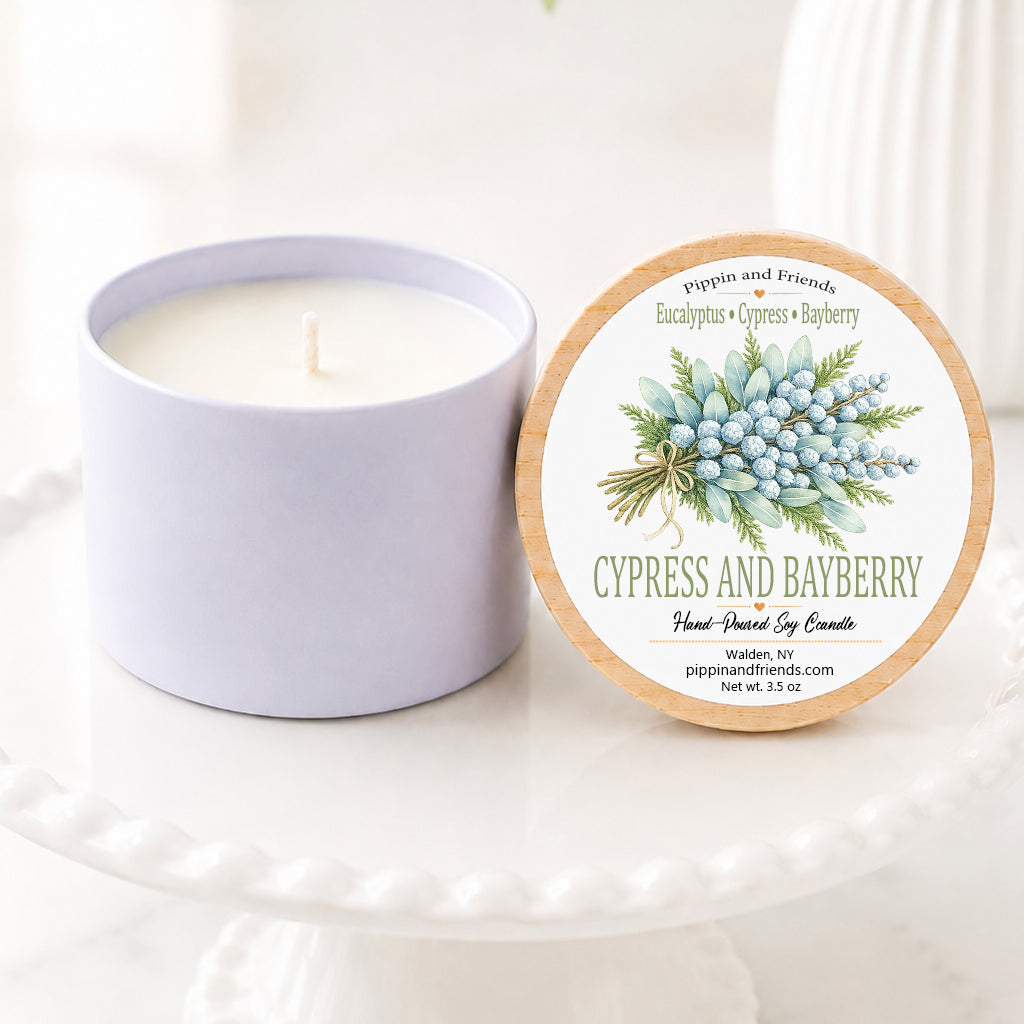 A soy candle jar sits beside its round lid on a white pedestal. The lid features a botanical label with blue berries and green leaves, reading 'Cypress and Bayberry' and 'Pippin and Friends.