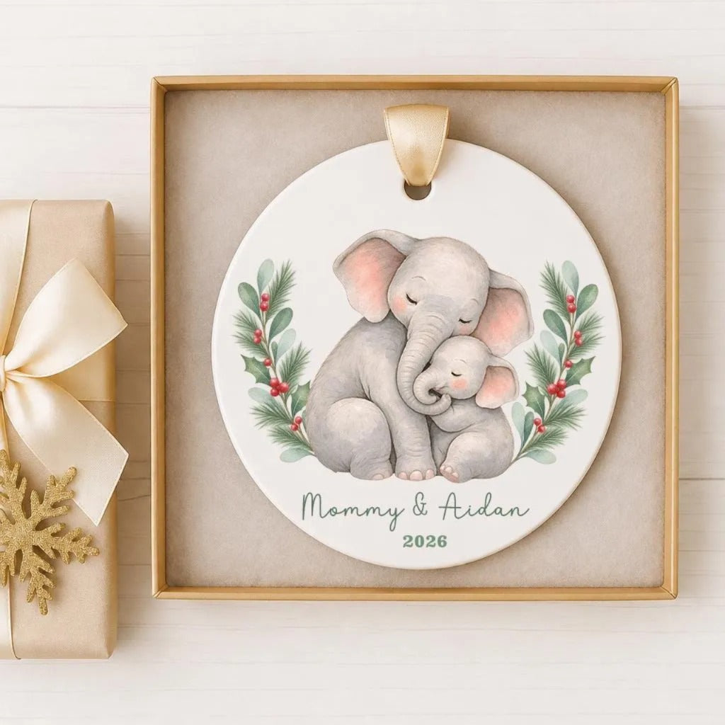 A ceramic ornament featuring a mother elephant and her baby, with the text "Mommy & Aidan" and the year "2026" displayed on it. The ornament is placed in a wooden frame and is accompanied by a gift box with a gold bow.