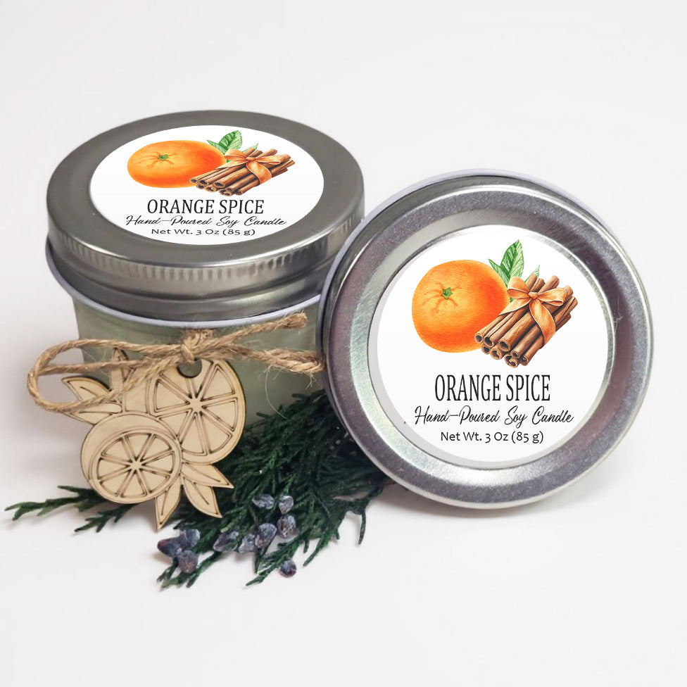 two jars of orange spice soy candles, with one jar placed on top of the other. The jars are decorated with orange and cinnamon sticks, and there are some pine branches and blueberries in the foreground.