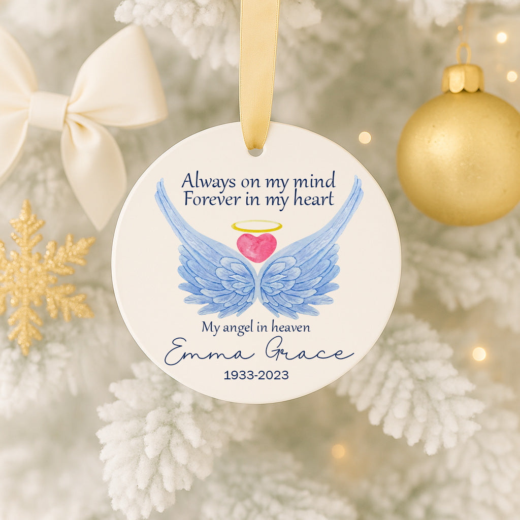 A white ornament with blue wings and a pink heart, hanging on a Christmas tree. The ornament has a message that reads "Always on my mind Forever in my heart" and "My angel in heaven Emma Grace 1933-2023".