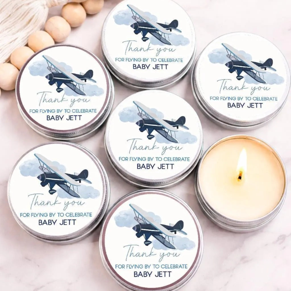 Personalized airplane baby shower candle favors in silver tins featuring a vintage blue airplane and cloud design, displayed on a marble surface with one candle lit.