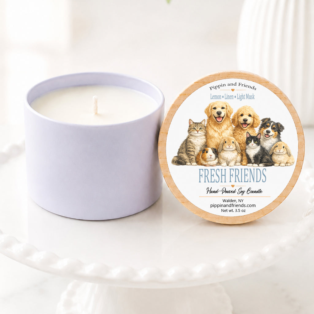 A soy candle in a white jar sits on a white pedestal next to its round lid, which features an illustration of various pets and the text 'Fresh Friends. The background is a bright, softly blurred indoor setting with a white vase of flowers.