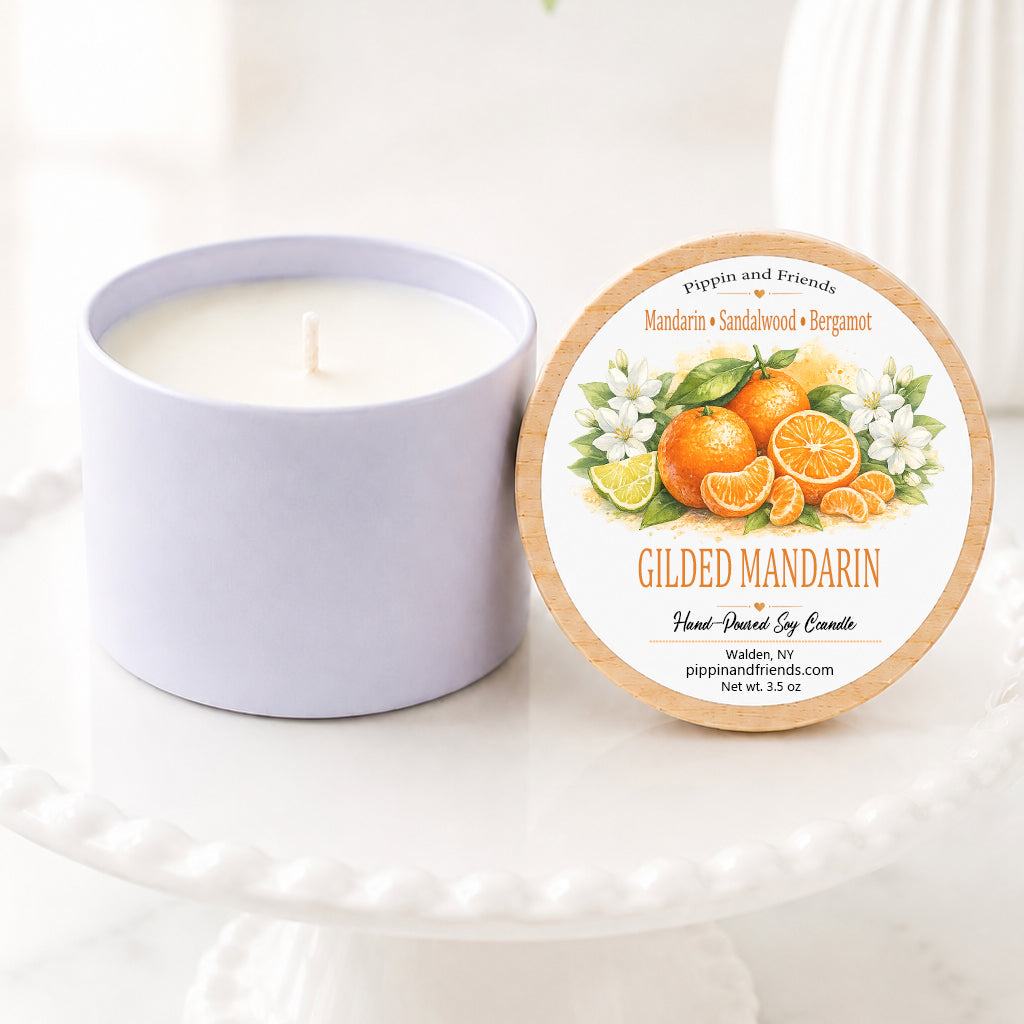 A soy candle jar next to its round lid, which features an illustration of gilded mandarins and white flowers. The candle and lid are displayed on a white ceramic pedestal stand with a soft-focus background of green leaves and white flowers.
