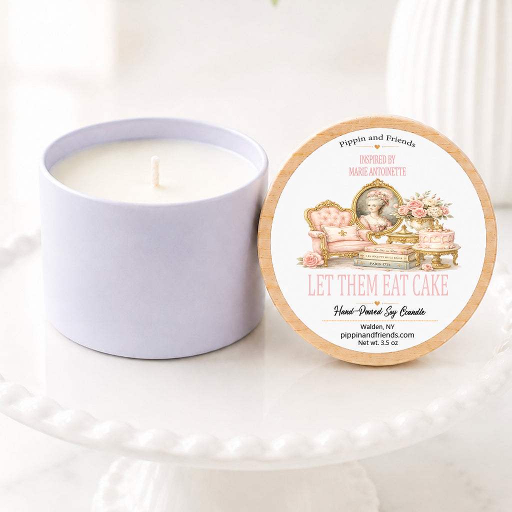 A soy candle sits on a white scalloped pedestal beside its matching wooden lid. The lid features an elegant Marie Antoinette-inspired label with a watercolor illustration of a pink Rococo chair, gilded portrait, roses, ornate cake, and antique books. The label reads "Inspired by Marie Antoinette – Let Them Eat Cake" with "Hand-Poured Soy Candle" below. The scene is styled on a bright marble surface with a softly blurred white vase of flowers in the background, creating a light, airy, French-