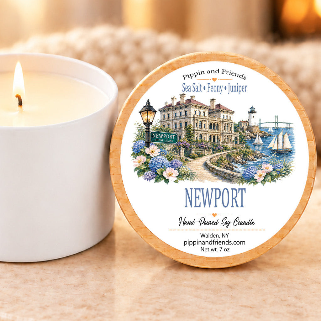 A round tin of Newport hand-poured soy candle sits beside a lit white candle jar on a stone surface. The tin label features an illustrated harbor scene with the text 'Pipkin and Friends' and 'Newport Hand-Poured Soy Candle.