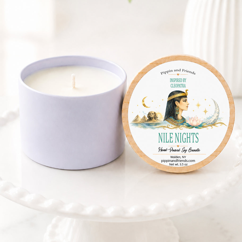 A hand-poured soy candle sits on a white scalloped pedestal beside its matching wooden lid. The lid features an elegant Cleopatra-inspired label with a watercolor illustration of Cleopatra in profile, the Egyptian pyramids and Sphinx, a crescent moon, lotus flower, and shimmering Nile waters. The label reads "Inspired by Cleopatra – Nile Nights" with "Hand-Poured Soy Candle" below. The candle is styled on a bright marble surface with a softly blurred white vase of flowers in the background, cr