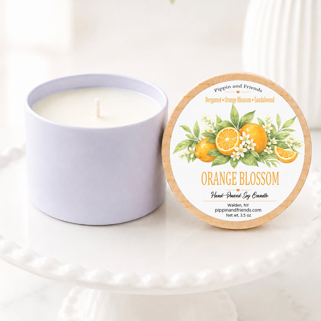 A soy candle in a matte jar sits next to its lid on a white pedestal. The lid features an illustration of oranges and the text 'ORANGE BLOSSOM' and 'pippin and friends'.