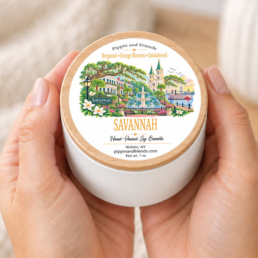 Hands holding a 7 oz Savannah soy candle with wood-look lid, featuring orange blossom, bergamot and sandalwood fragrance and illustrated Savannah destination label.