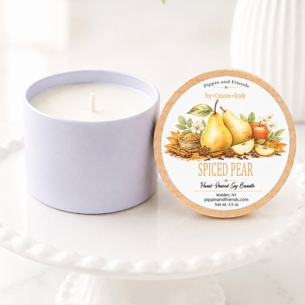 A soy candle next to its round lid, which features an illustration of spiced pears. The candle and lid are displayed on a white ceramic pedestal stand with a soft-focus background of green leaves and white flowers.