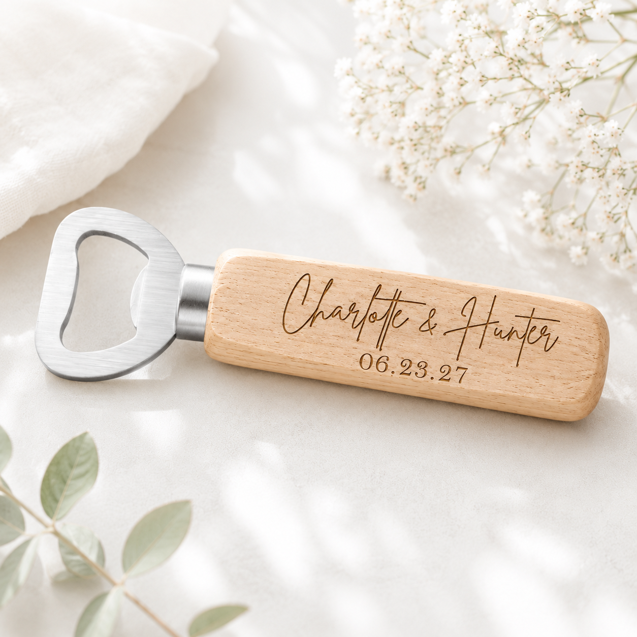 Personalized wooden bottle opener with custom engraved names and wedding date on a natural wood handle with stainless steel opener, styled in a light and airy wedding-inspired setting.