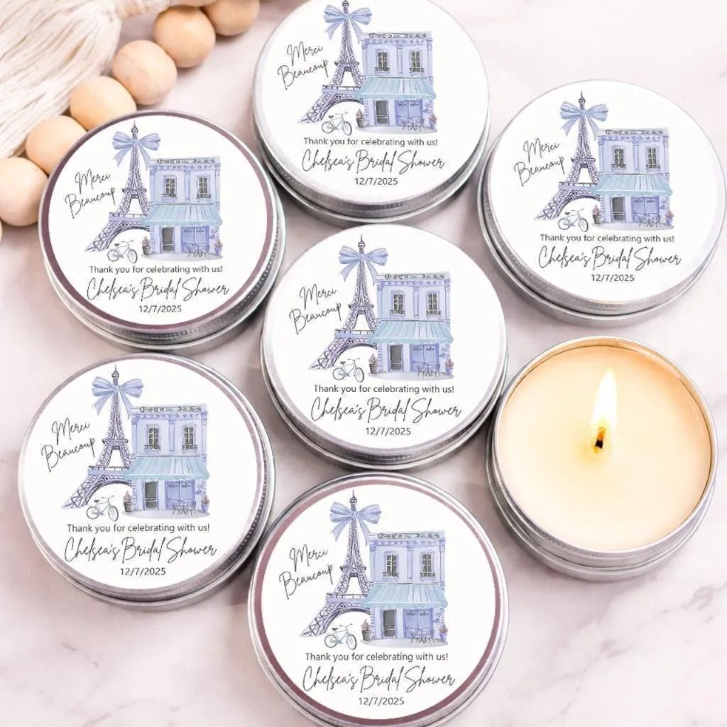 Personalized Paris bridal shower candle favors in silver tins featuring a watercolor Eiffel Tower, Paris café, and custom event details, displayed on a marble surface with one candle lit.