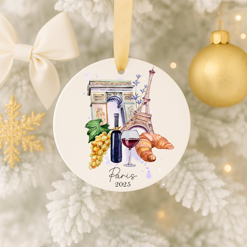A decorative ornament featuring a Paris-themed design, including the Eiffel Tower, grapes, a wine glass, and a croissant, hanging on a Christmas tree.