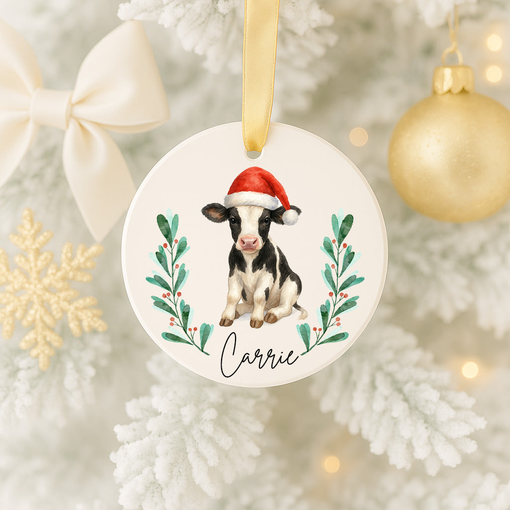 A white Christmas tree ornament with a cow wearing a Santa hat and the name "Carrie" written on it.