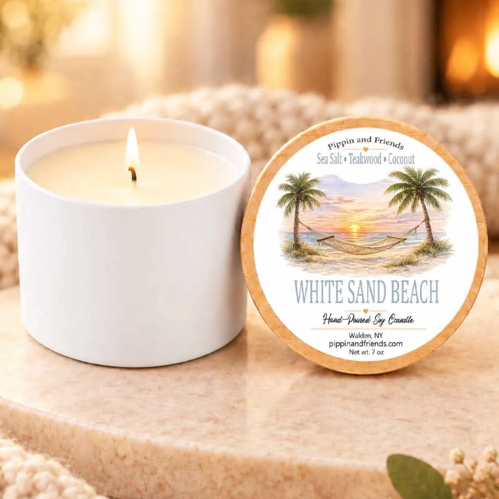 a white candle labeled "White Sand Beach" next to a white candle holder, both sitting on a wooden surface.