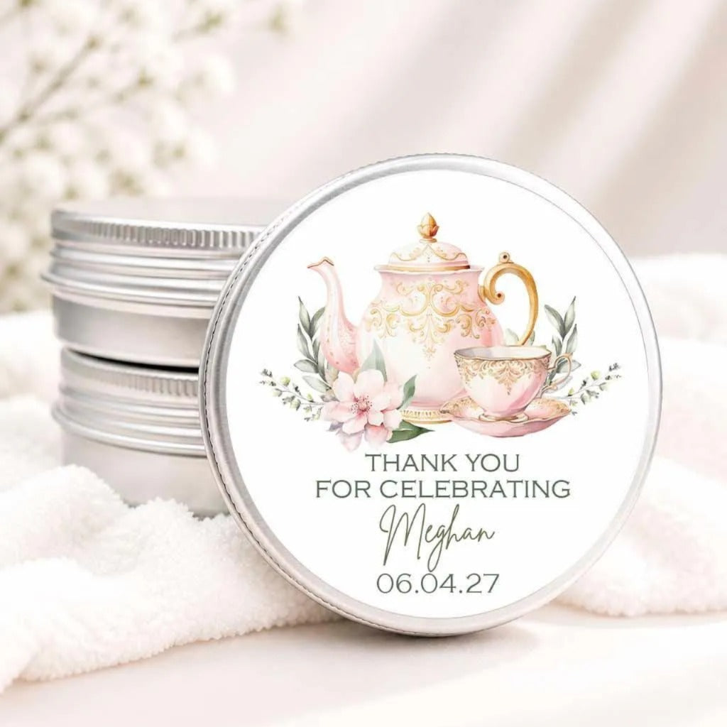 A round tin lid with a decorative teapot and flowers on it, placed on a white cloth. The lid has a message that says "THANK YOU FOR CELEBRATING Meghan 06.04.27".