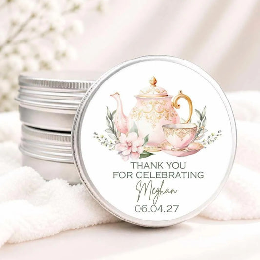 A round tin lid with a decorative teapot and flowers on it, placed on a white cloth. The lid has a message that says "THANK YOU FOR CELEBRATING Meghan 06.04.27".
