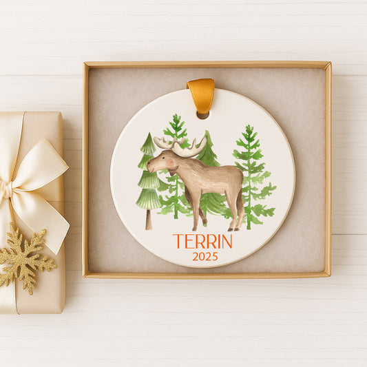 A white ceramic ornament with a moose and pine trees, displayed in a gold box with a gold ribbon.