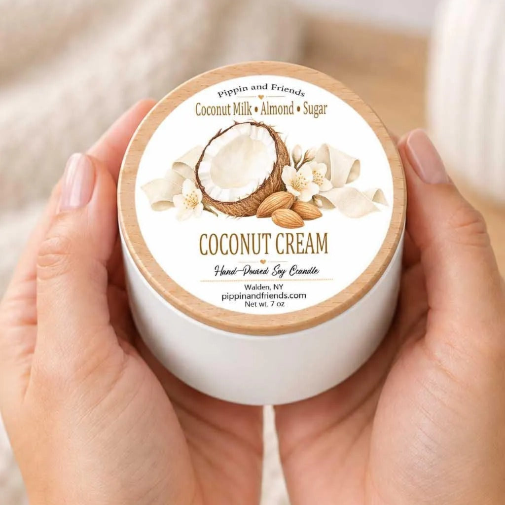 A person's hands holding a white container with a label that features an illustration of a coconut and almonds, along with the text "COCONUT CREAM Hand-Dipped Soy Candle" and "pippin and Friends".