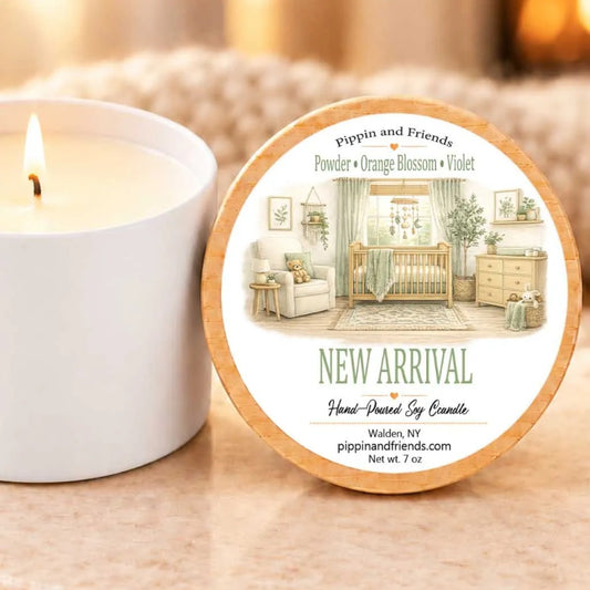 a white candle with a label that reads "New Arrival Hand-Poured by Candle" and "Pippin and Friends Powder + Orange Blossom + Violet". The candle is placed next to a white candle holder on a table.