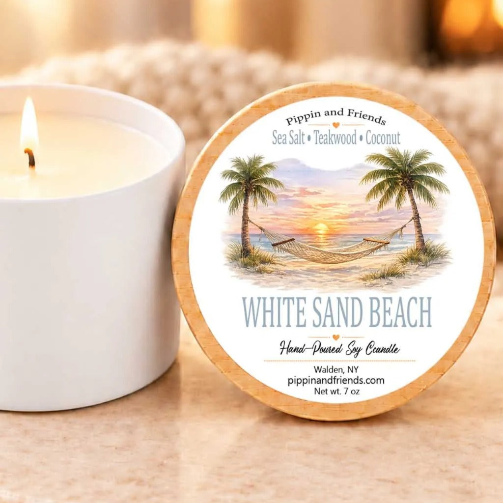 a white candle with a label that reads "White Sand Beach" and "Hand-poured by Candle" on a wooden coaster. The candle is placed next to a white candle holder, and there is a blurred background that includes a fireplace.