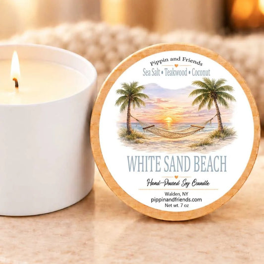 a white candle with a label that reads "White Sand Beach" and "Hand-poured by Candle" on a wooden coaster. The candle is placed next to a white candle holder, and there is a blurred background that includes a fireplace.