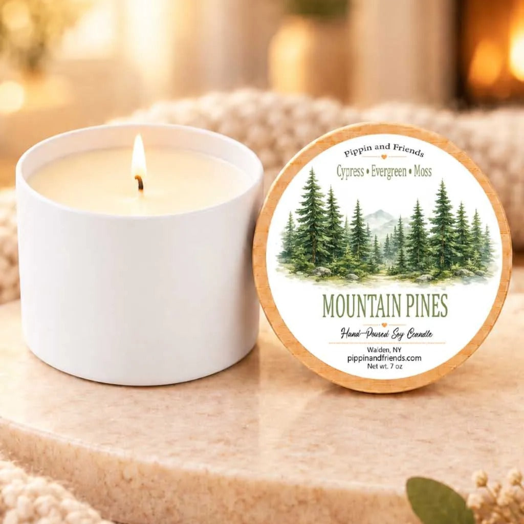 a white candle labeled "Mountain Pines" next to a white candle, both sitting on a wooden surface.
