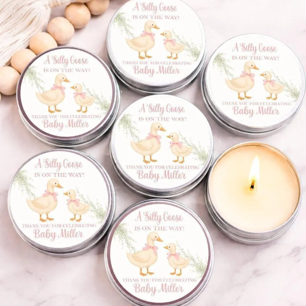 a collection of round tin candles with illustrations of two baby geese, along with text that reads "A Silly Goose is on the way! Thank you for celebrating Baby Miller". The candles are arranged on a marble surface, with some of them lit, creating a warm and cozy atmosphere.